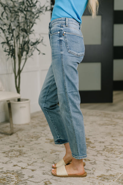 A person wearing light-wash blue cropped jeans, a bright blue ribbed top, and beige slide sandals, standing on a patterned rug in an indoor setting.