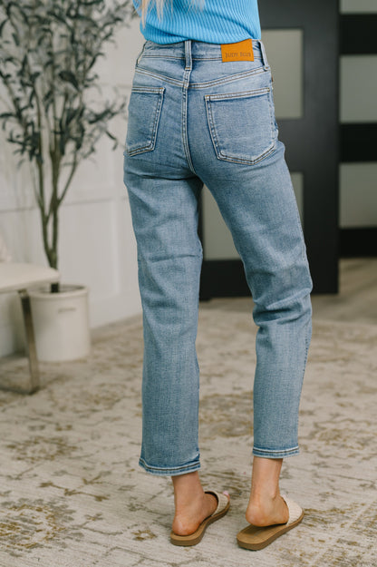 A person shown from the back wearing light-wash blue cropped jeans with a bright orange brand patch on the waistband, standing indoors on a patterned rug.