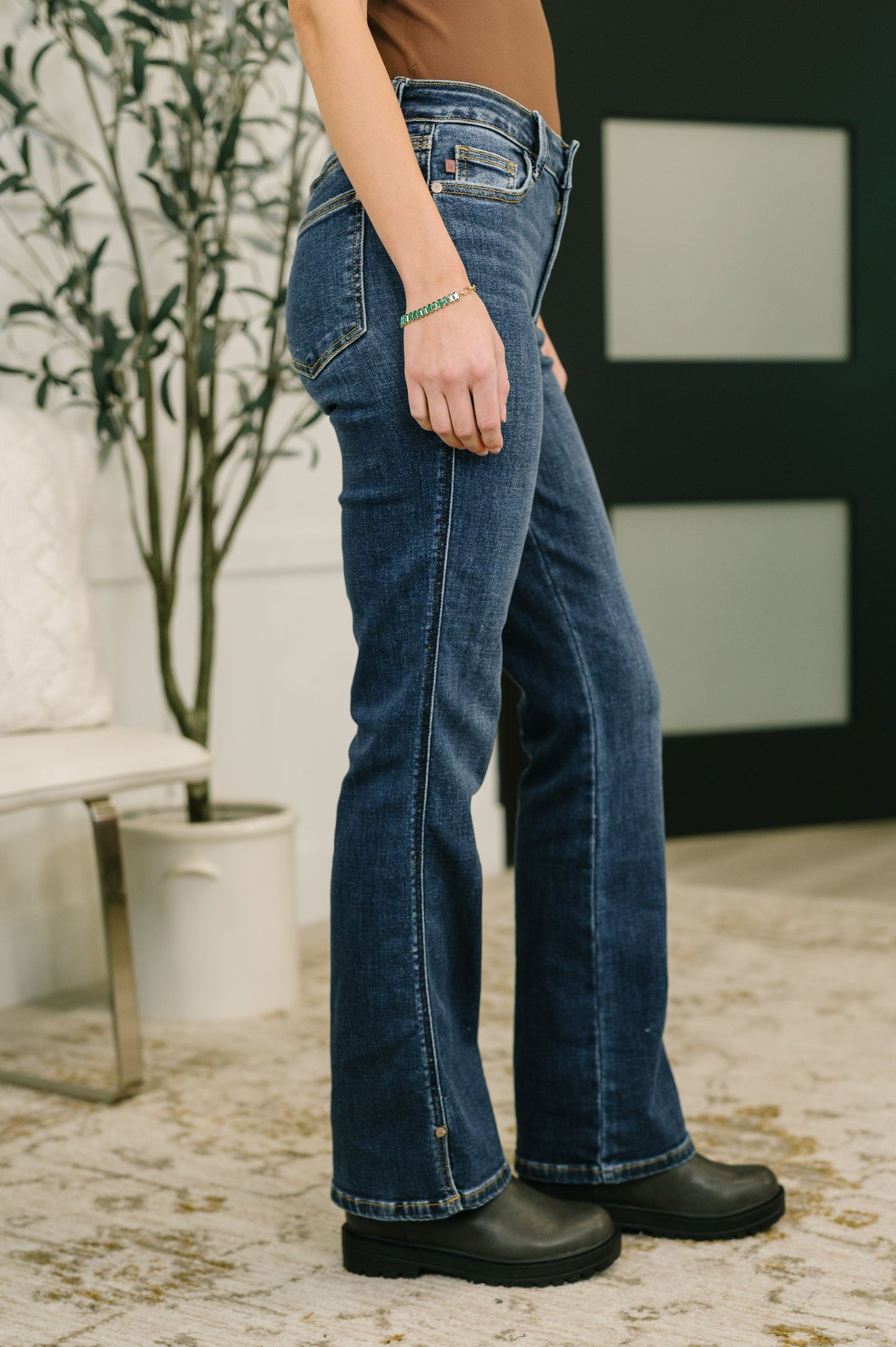 Person wearing blue jeans and black boots standing indoors with a plant and white wall in the background