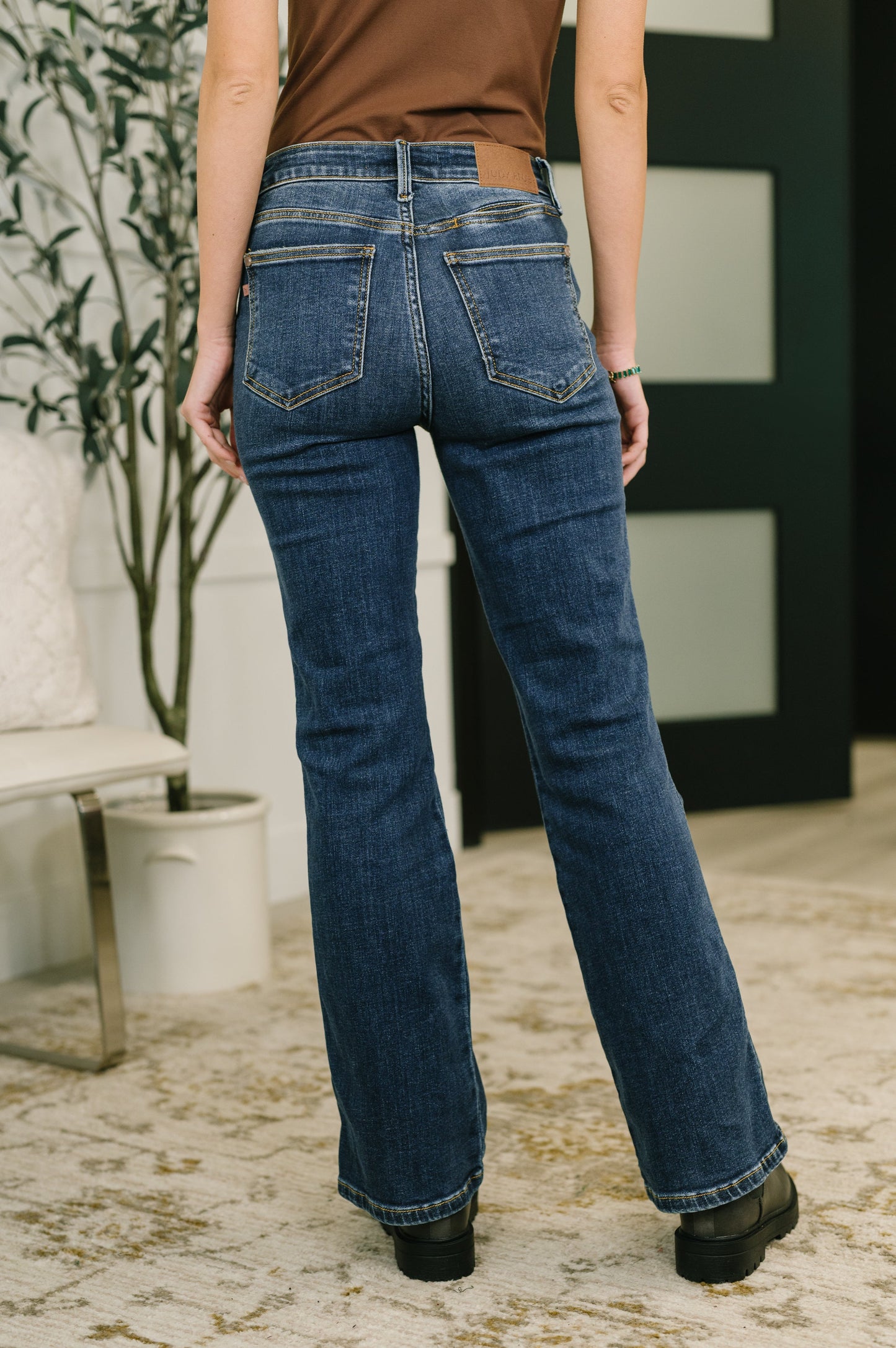 Person wearing blue jeans standing in a room with a plant and white wall. Judy Blue