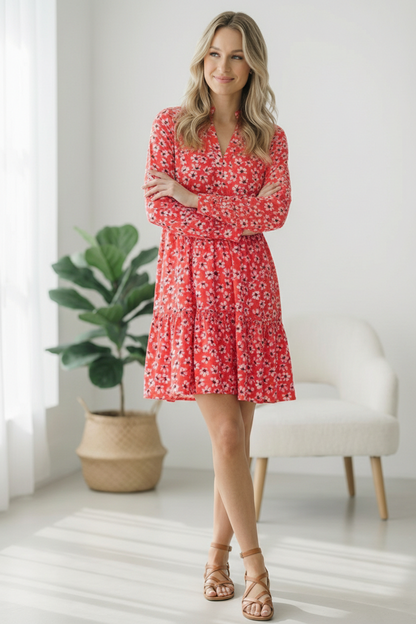 Woman wearing a red floral dress standing in a bright room with a plant and chair.