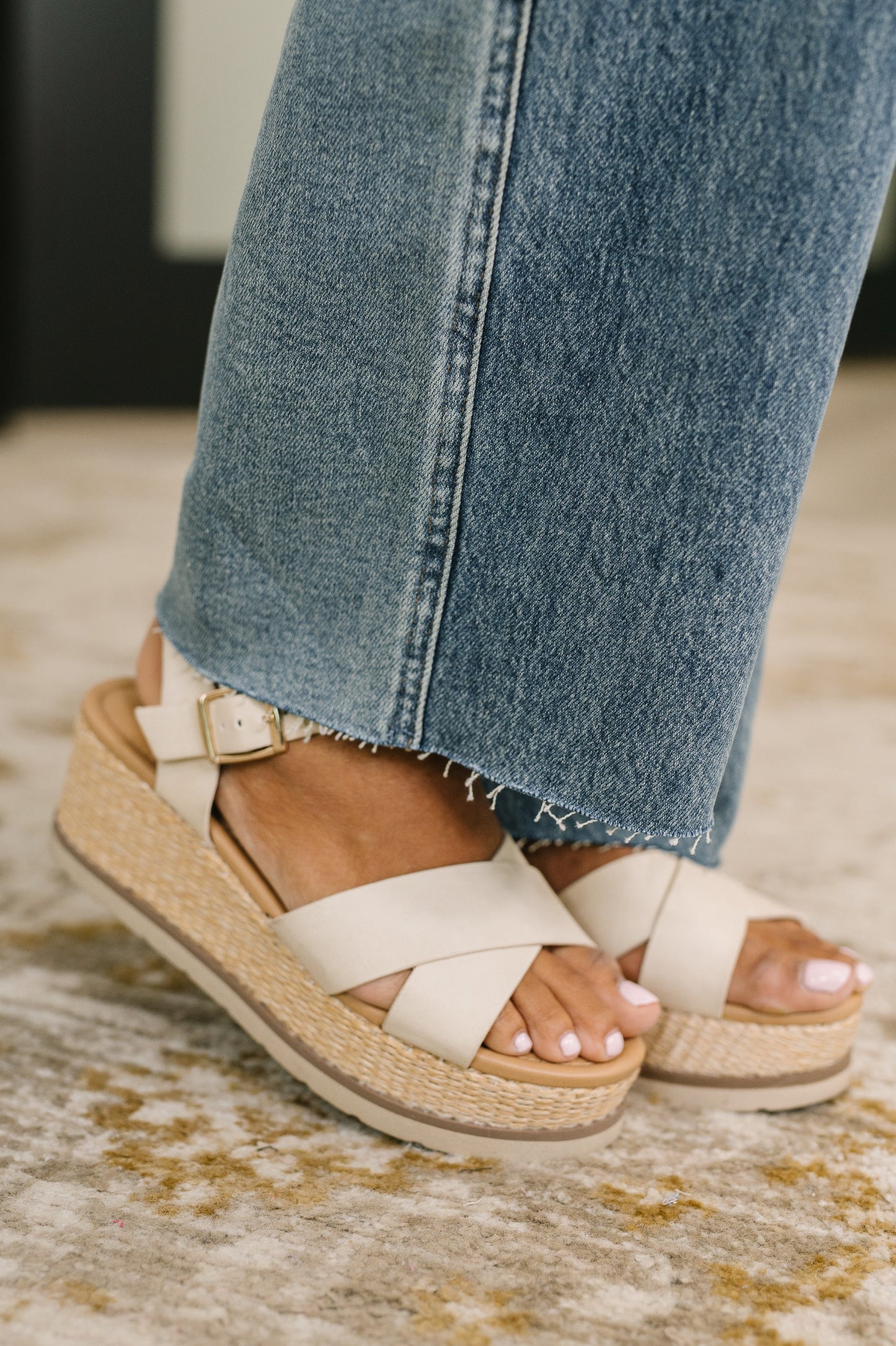 Beige sandals with wicker sole worn with blue jeans on a patterned rug.