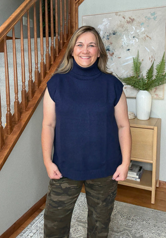 Woman wearing a navy sleeveless sweater and camouflage pants standing in a home setting.