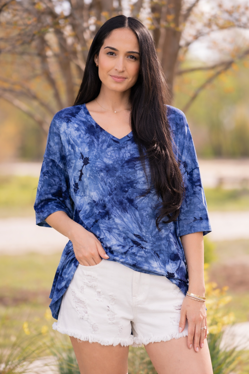 Woman wearing a blue tie-dye shirt and white shorts standing outdoors with trees in the background