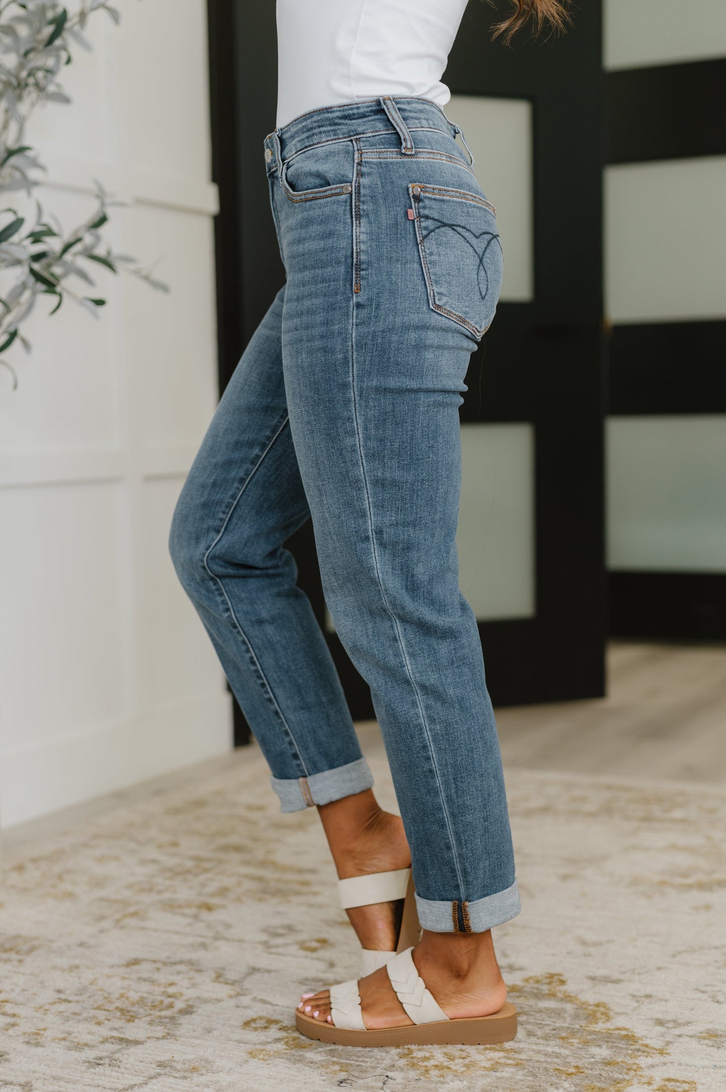 A person wearing light blue cuffed denim jeans with a white top and white strappy flat sandals, standing indoors on a patterned rug.