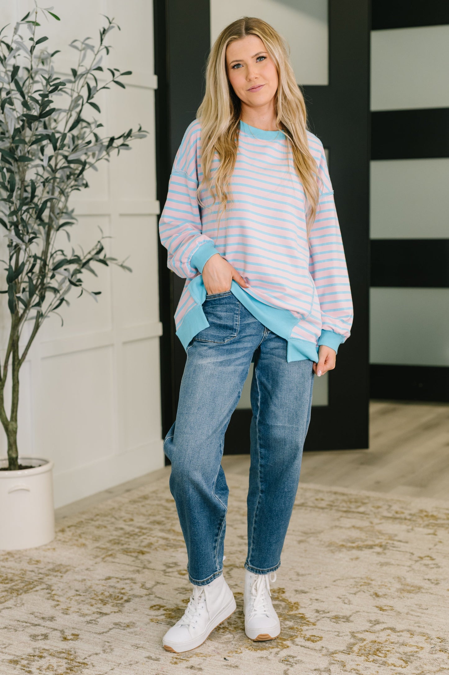 Woman wearing a striped sweater and jeans standing indoors with a plant in the background