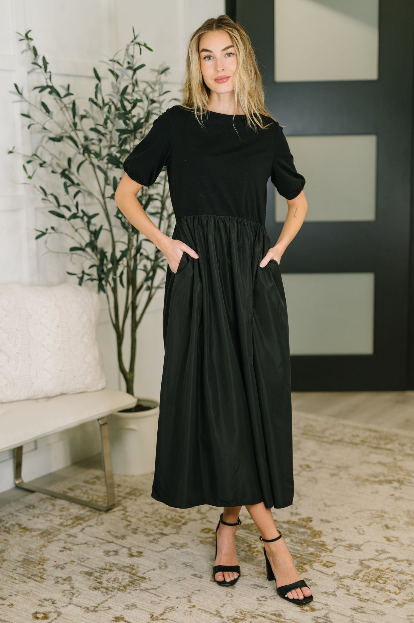 Woman wearing a black dress standing in a room with a plant and bench.