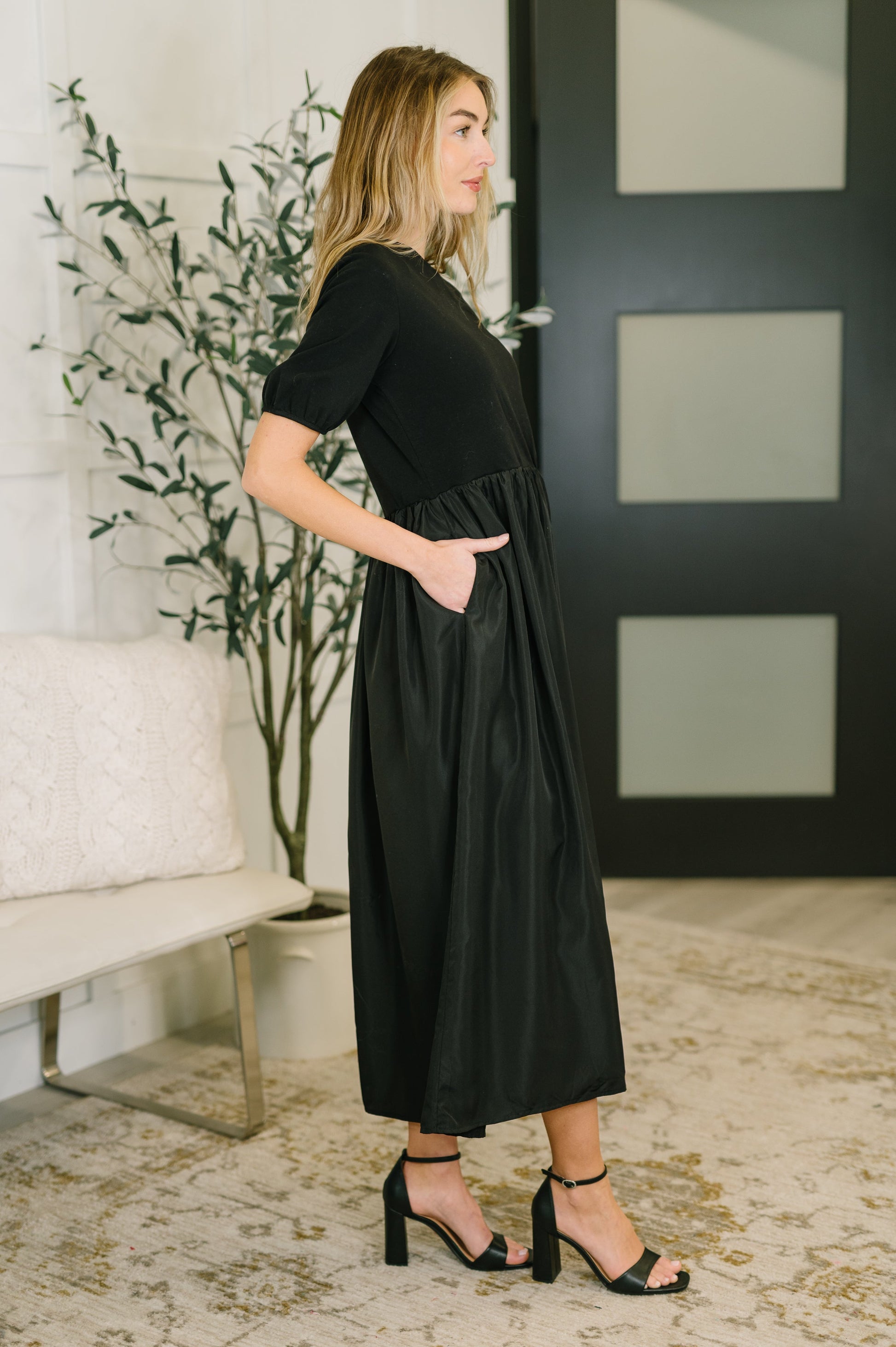 Woman wearing a black dress standing in a room with a plant and a bench.