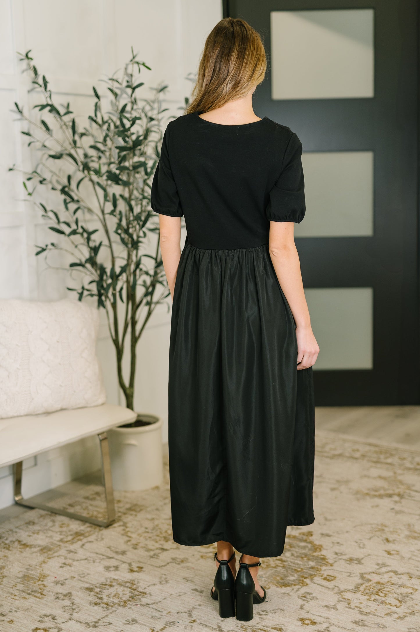 Woman wearing a black dress standing in a room with a plant and wall art.