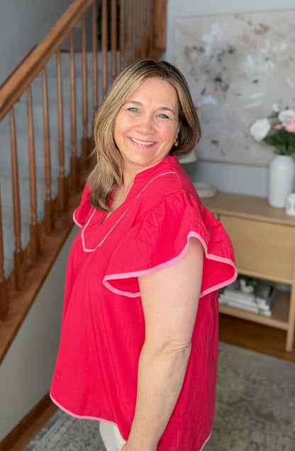 Woman in a red blouse standing in a home setting with a staircase and decorative items in the background.
