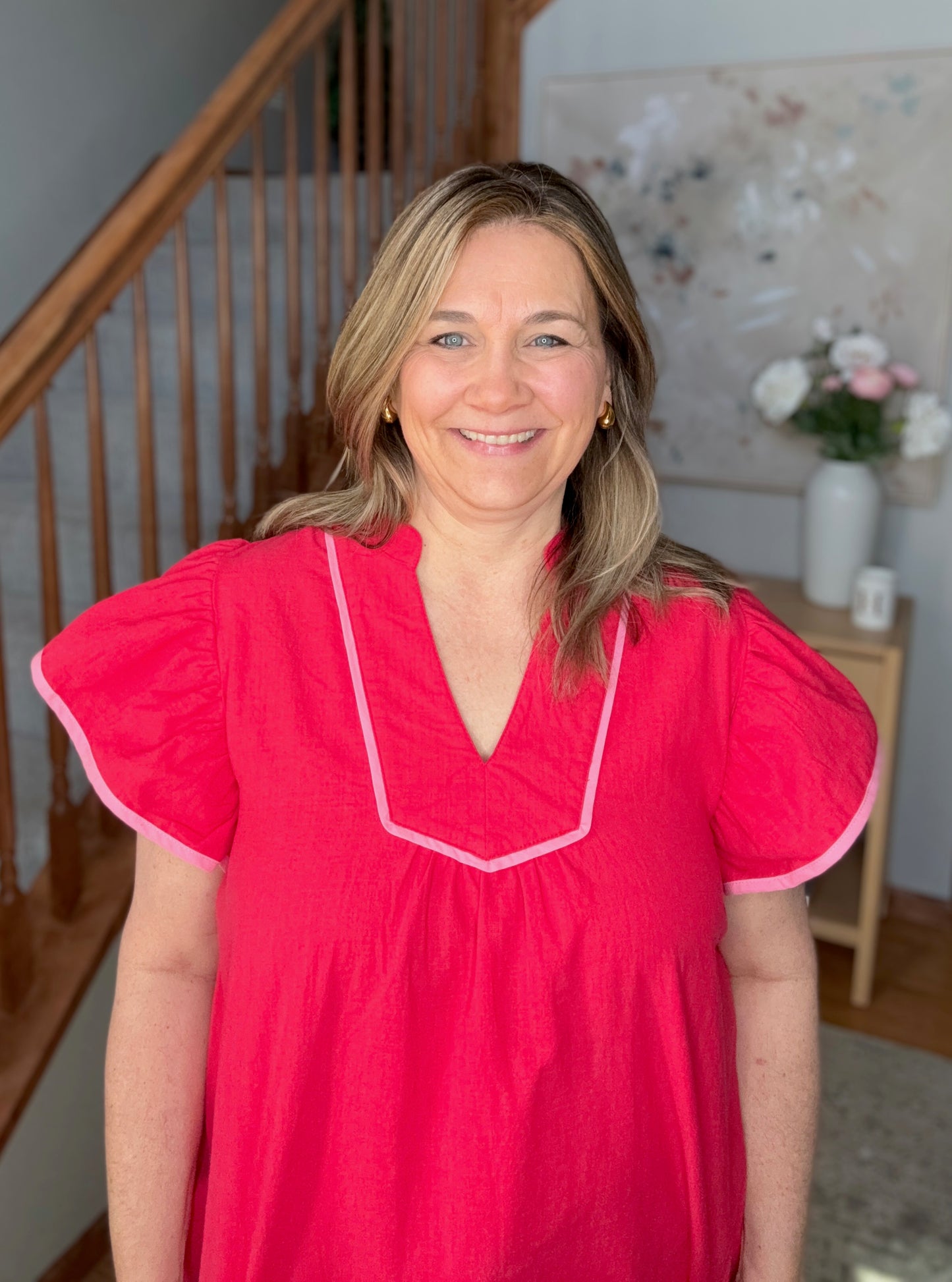 Woman wearing a pink blouse standing in a home setting with a staircase and flowers in the background.