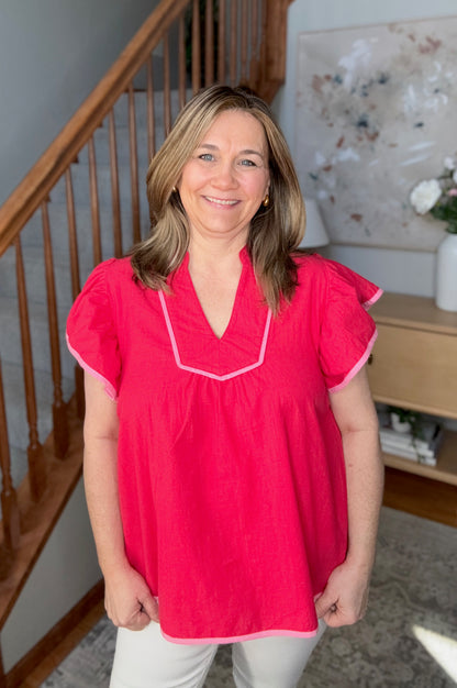 Woman wearing a pink blouse standing in a home setting with a staircase and decor in the background.