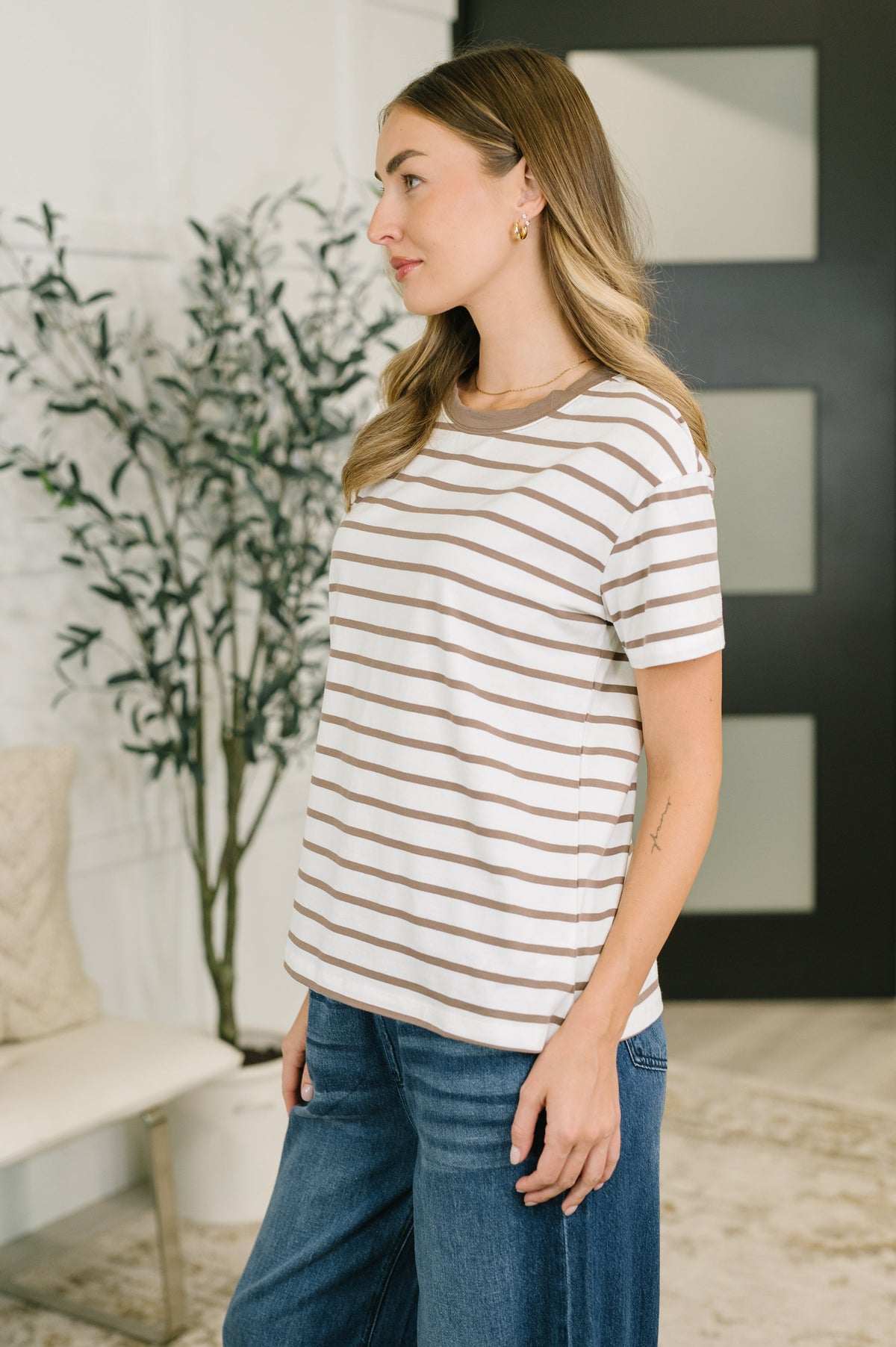 A woman wearing a short-sleeve crewneck top with horizontal beige and white stripes, paired with blue jeans, standing in a modern indoor setting.