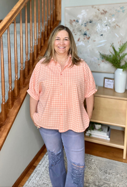 Woman standing in a home interior wearing a pink checkered shirt and blue jeans.