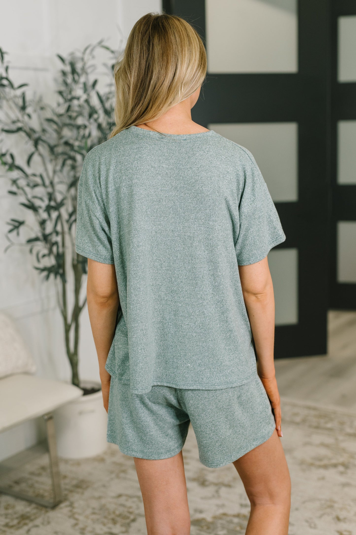 A woman shown from behind wearing a heathered sage green short-sleeve t-shirt and matching shorts in a modern indoor setting.