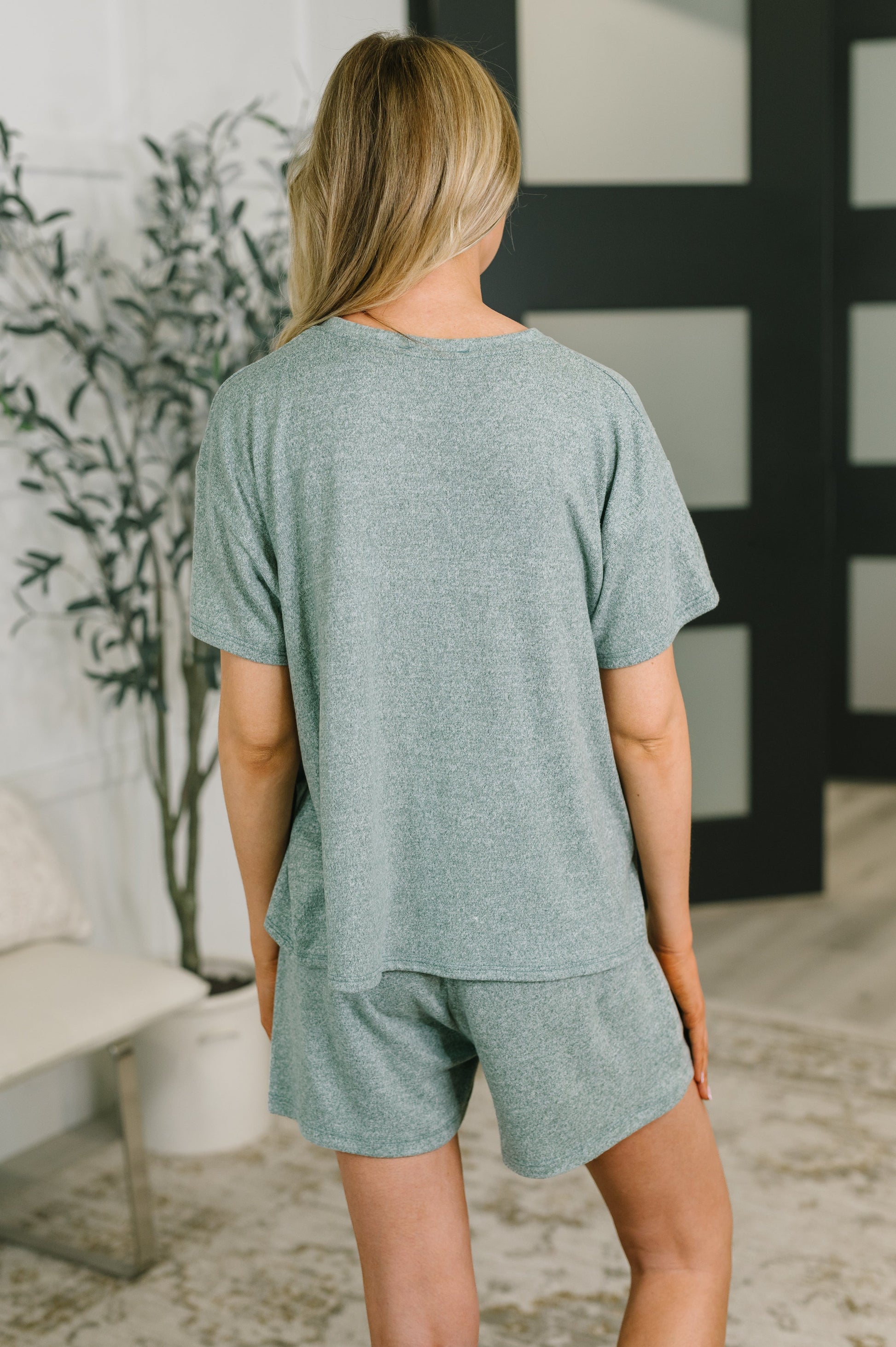 A woman shown from behind wearing a heathered sage green short-sleeve t-shirt and matching shorts in a modern indoor setting.