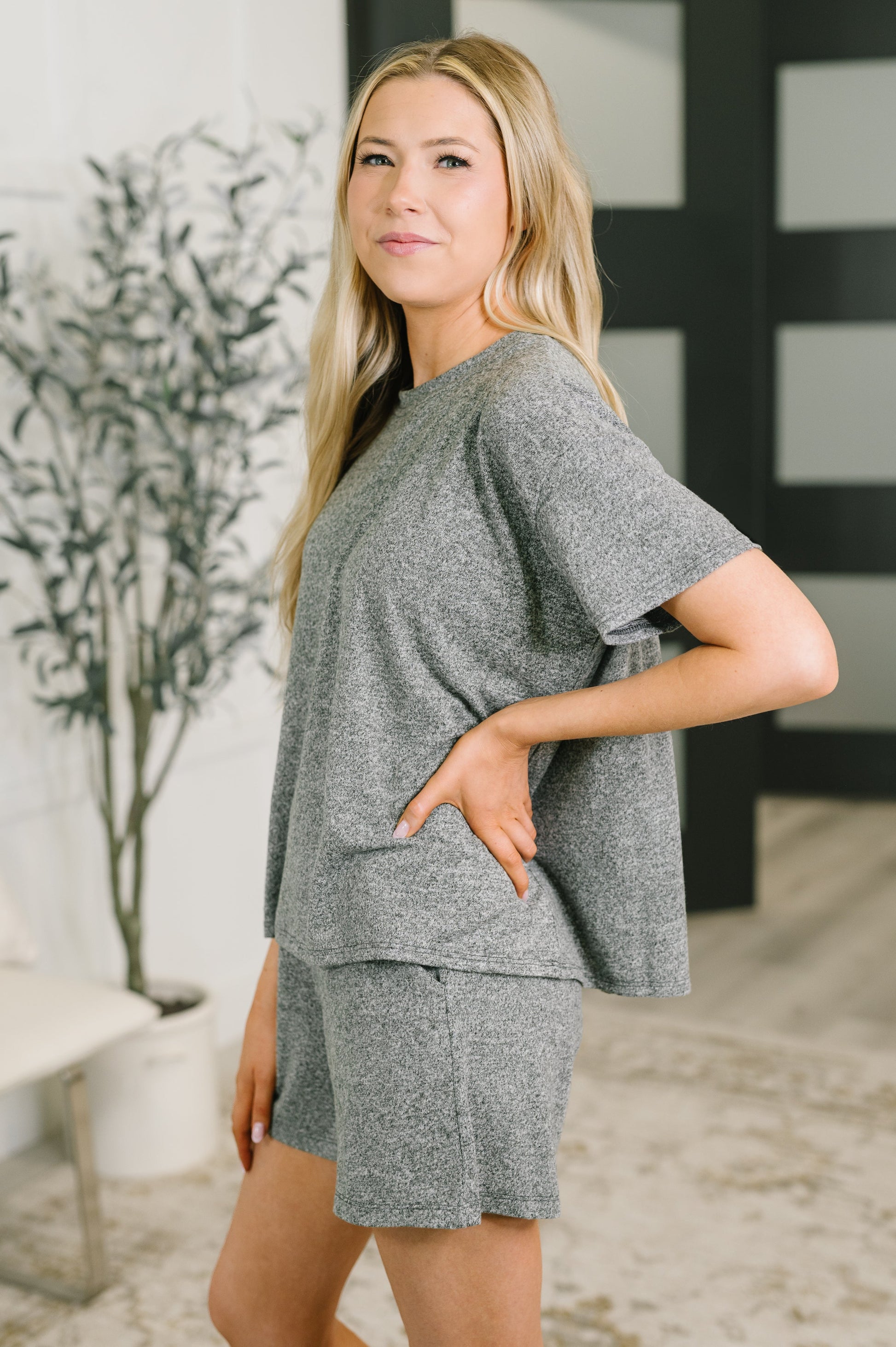 A woman modeling a heather gray two-piece lounge set consisting of a short-sleeve top and matching shorts, standing in a modern indoor setting.
