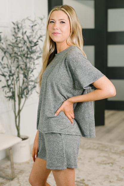 A woman modeling a heather gray two-piece lounge set consisting of a short-sleeve top and matching shorts, standing in a modern indoor setting.