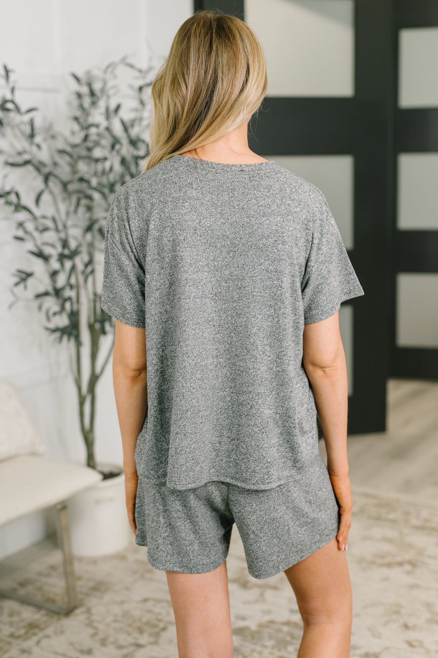 A woman viewed from behind wearing a heather gray short-sleeve t-shirt and matching shorts in a modern indoor setting.