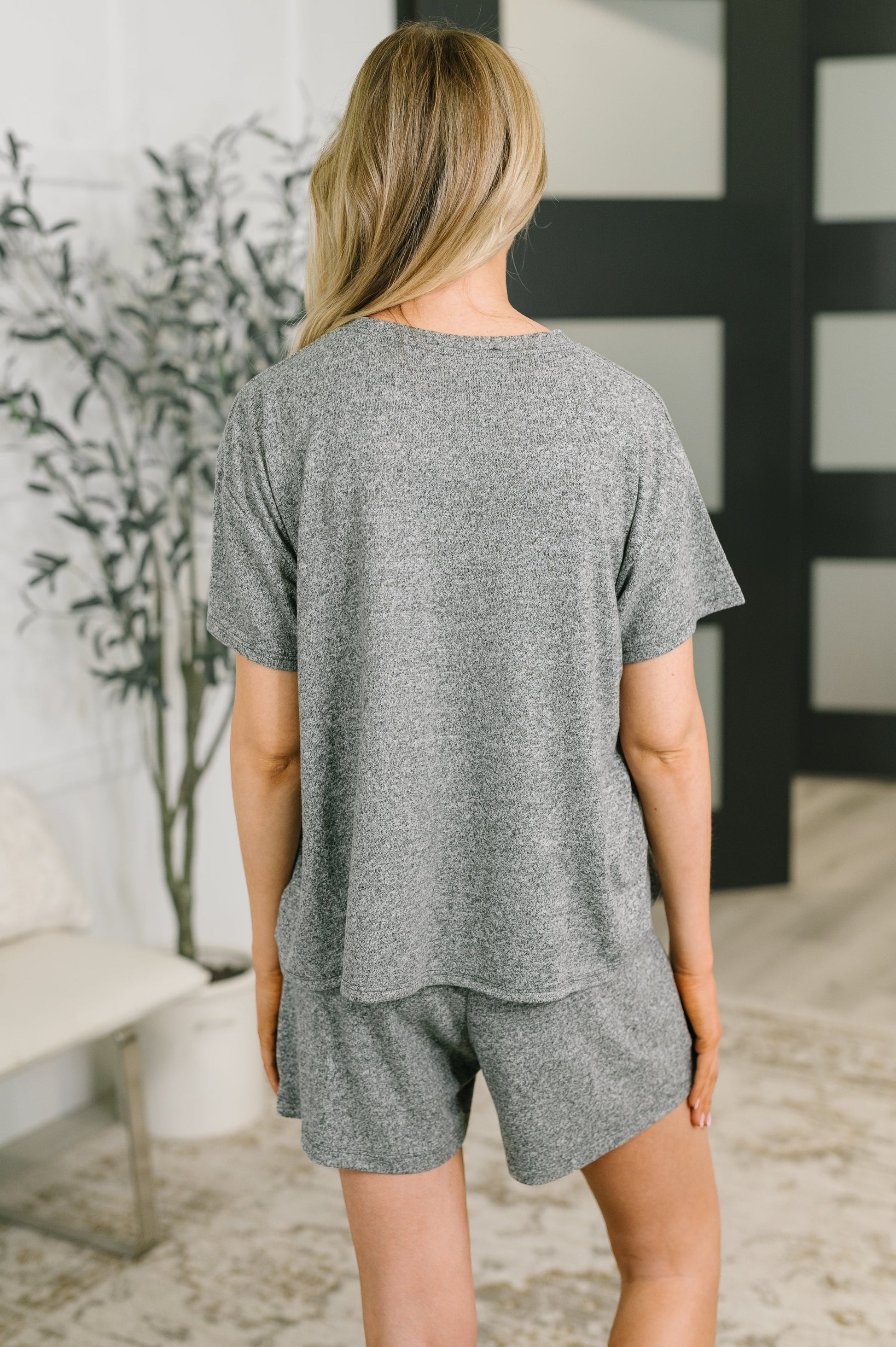 A woman viewed from behind wearing a heather gray short-sleeve t-shirt and matching shorts in a modern indoor setting.