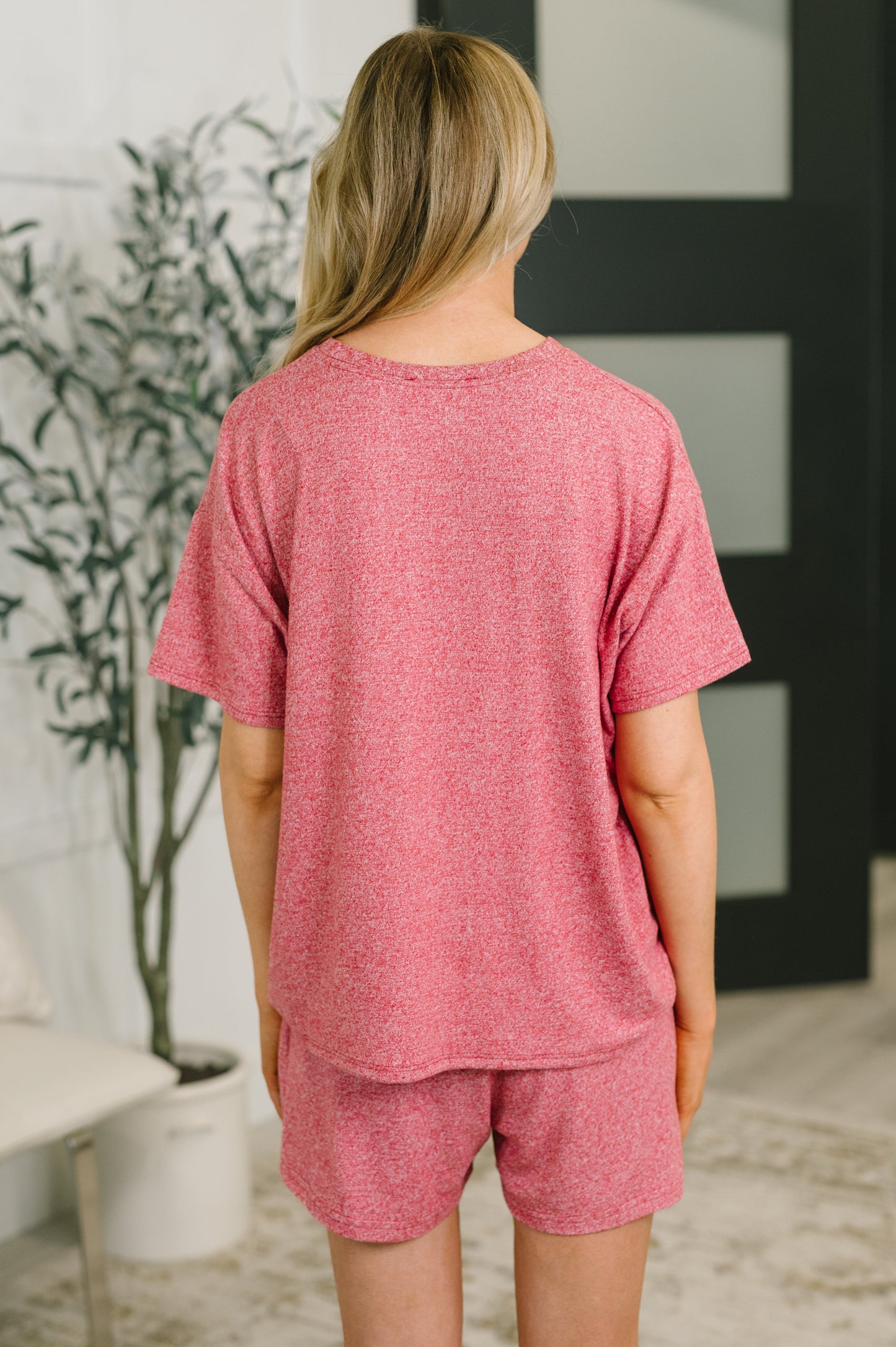 A woman viewed from behind wearing a heathered pink short-sleeve t-shirt and matching shorts set in a bright indoor room.