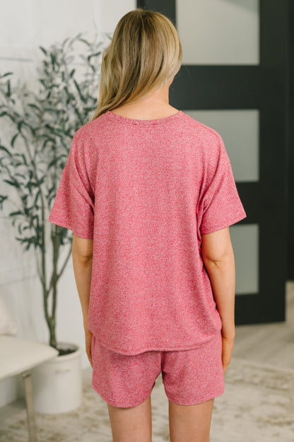 A woman viewed from behind wearing a heathered pink short-sleeve t-shirt and matching shorts set in a bright indoor room.