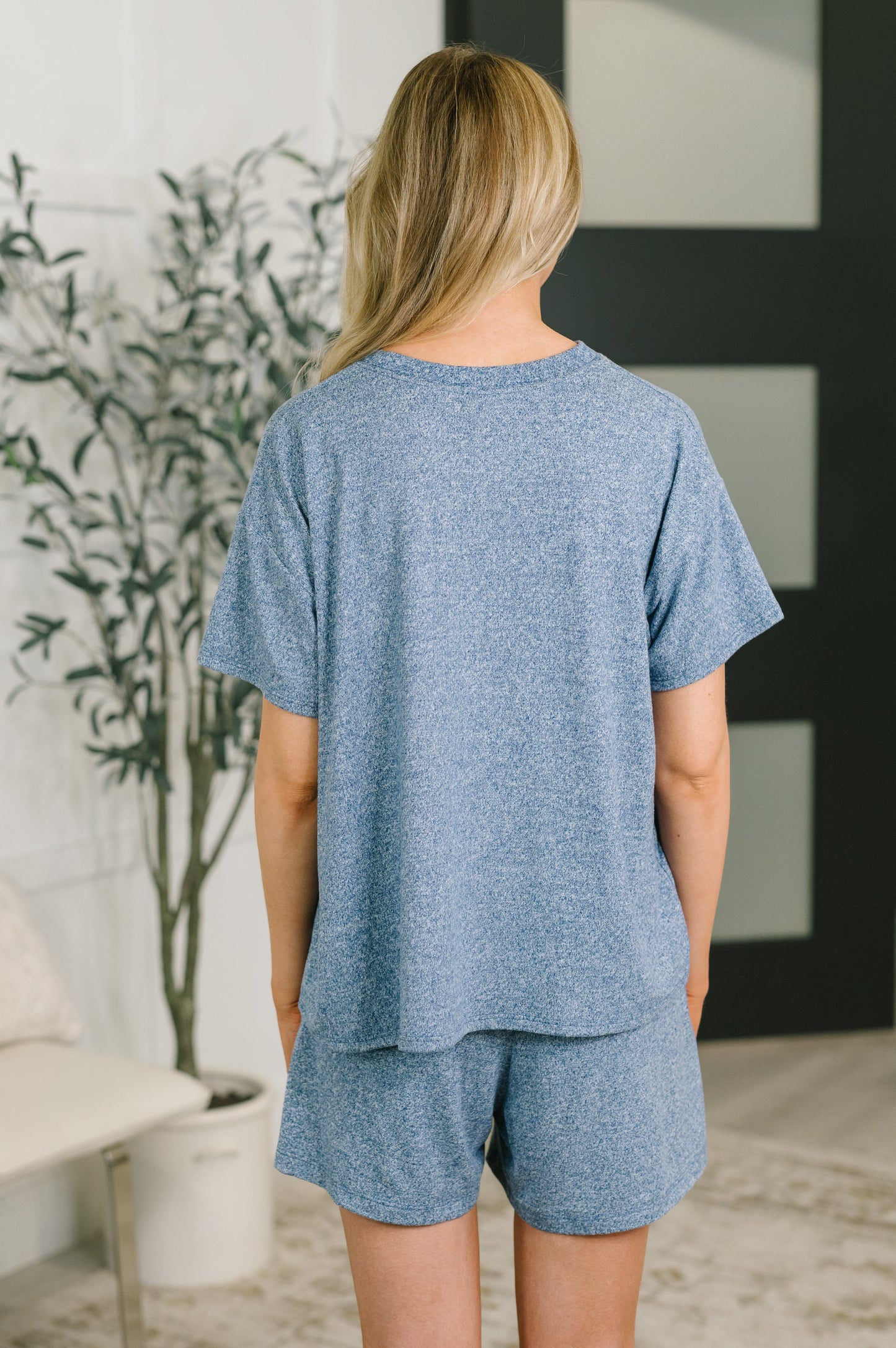 A woman viewed from behind wearing a heathered blue short-sleeve t-shirt and matching shorts set in a modern indoor room.