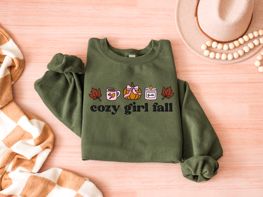 Green sweatshirt with 'cozy girl fall' text and autumn-themed graphics on a pink surface.
