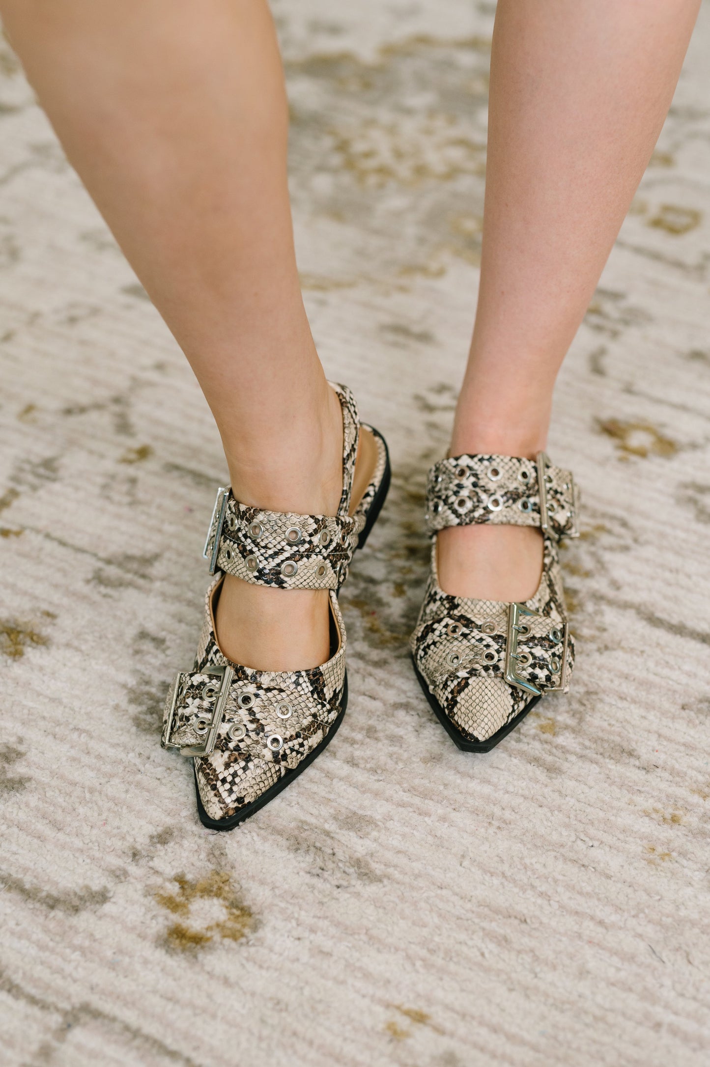 A close-up of a person's feet wearing pointed-toe Mary Jane shoes with a beige and black snakeskin pattern, standing on a textured beige rug.