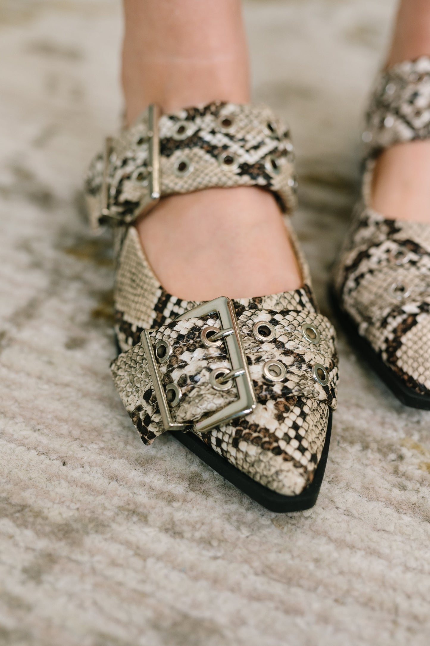 Close-up of a person wearing pointed-toe Mary Jane flats with a snakeskin pattern and large silver buckle straps, standing on a textured light rug.