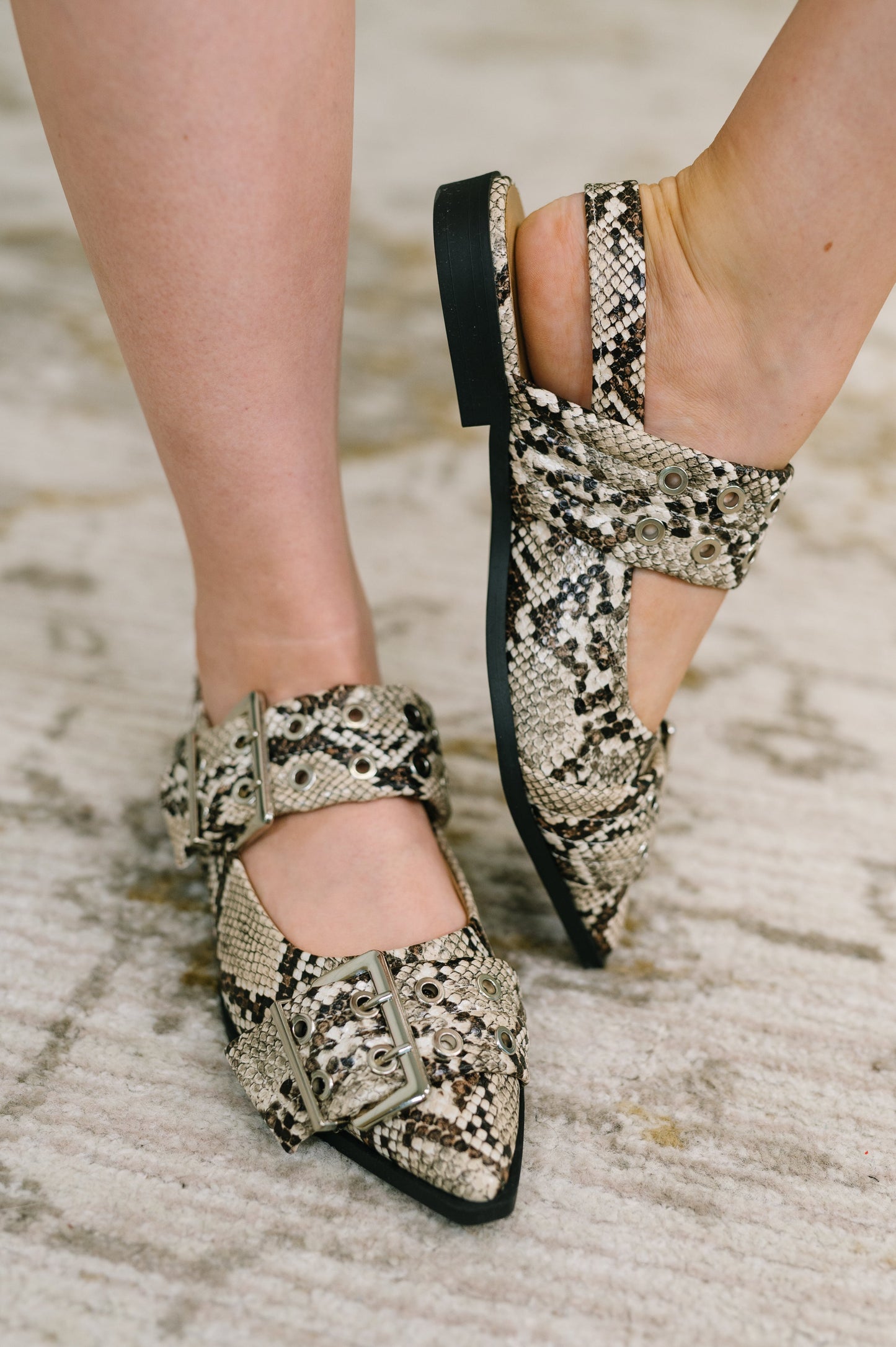 A close-up of a person's feet wearing pointed-toe flat mules with a beige, black, and brown snakeskin pattern, featuring double straps and square buckles, set against a neutral textured rug.