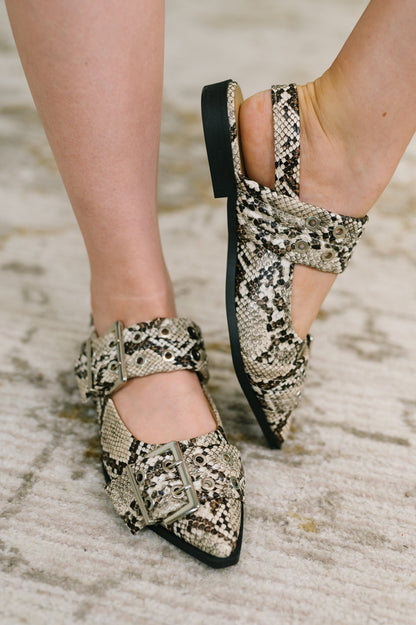 A close-up of a person's feet wearing pointed-toe flat mules with a beige, black, and brown snakeskin pattern, featuring double straps and square buckles, set against a neutral textured rug.