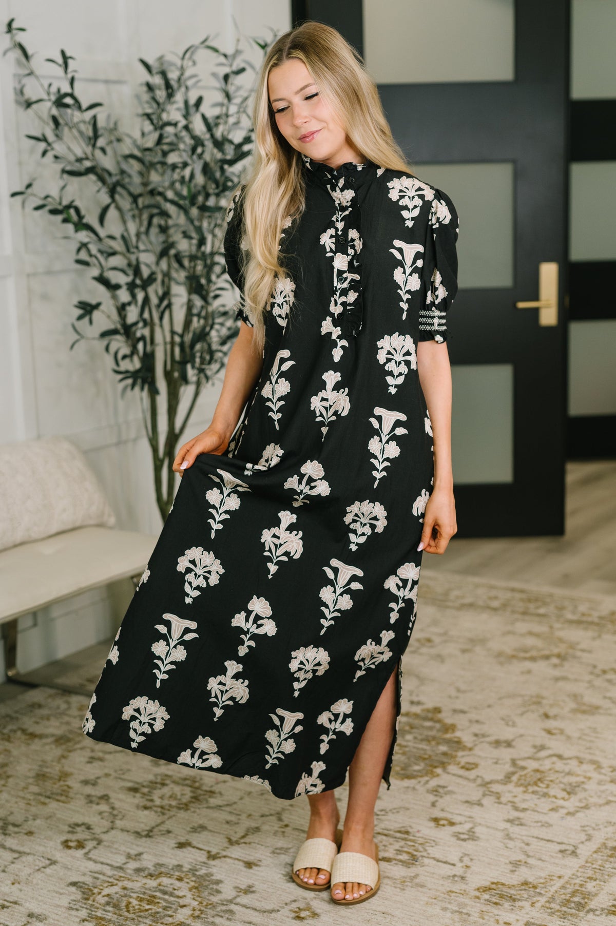 A woman modeling a black midi dress with a white floral pattern, standing in an indoor setting with a patterned rug, potted plant, and a dark door in the background.
