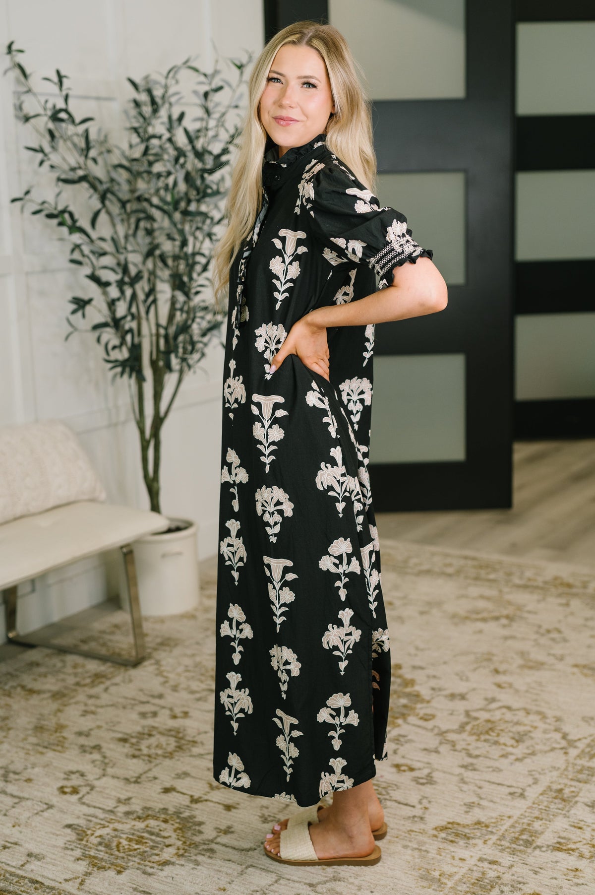 A woman modeling a black midi dress with a white floral pattern, standing in a modern indoor setting with a bench, potted plant, and patterned rug.