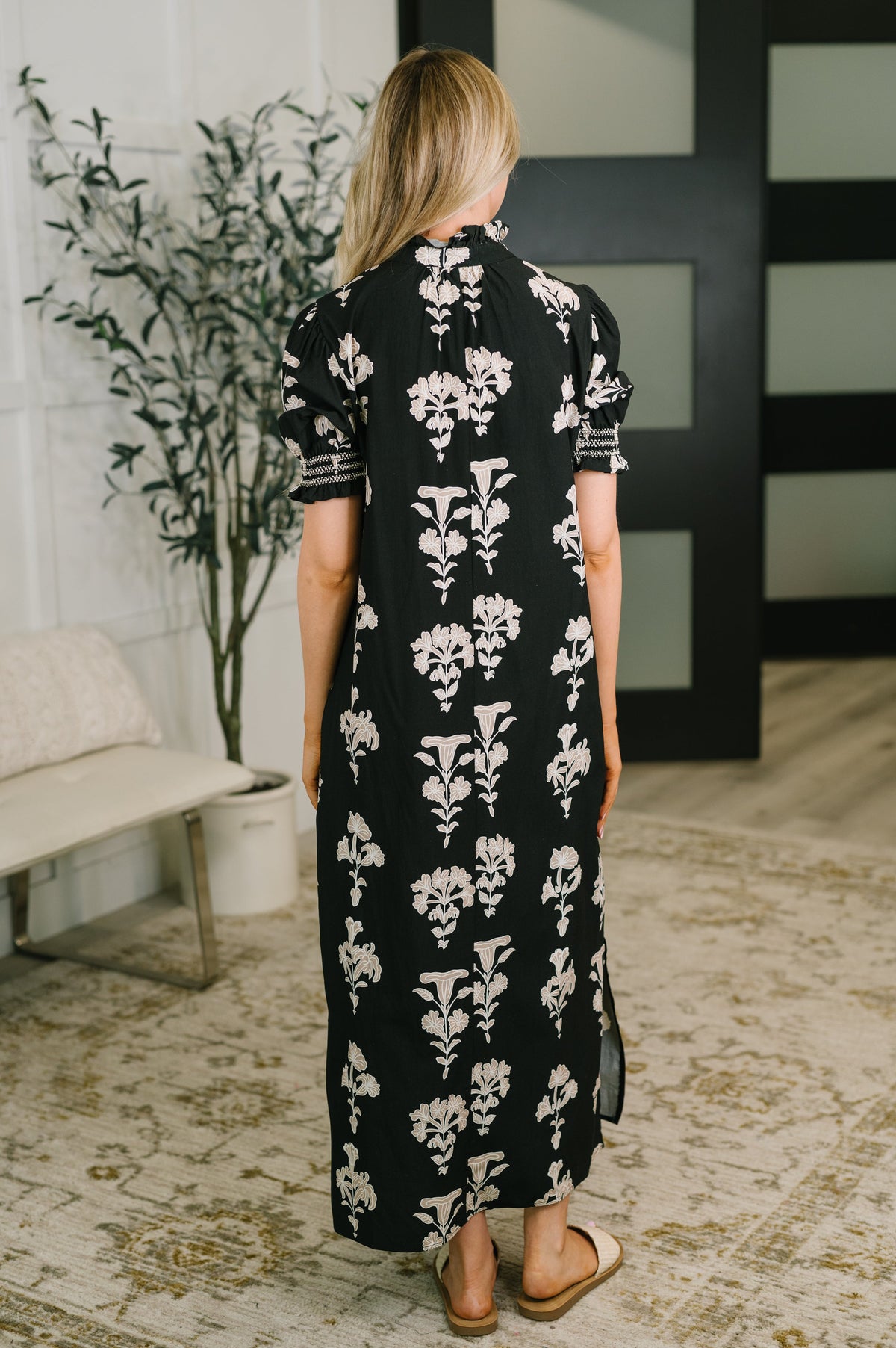 A woman viewed from behind wearing a black midi dress with a white floral pattern and short sleeves, standing in a modern indoor room with a bench, potted plant, and patterned rug.