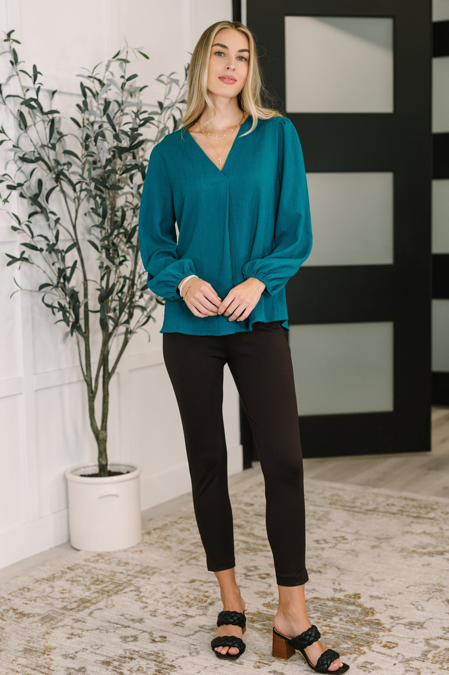 Woman wearing a teal blouse and black pants standing indoors with a plant and decorative wall in the background.