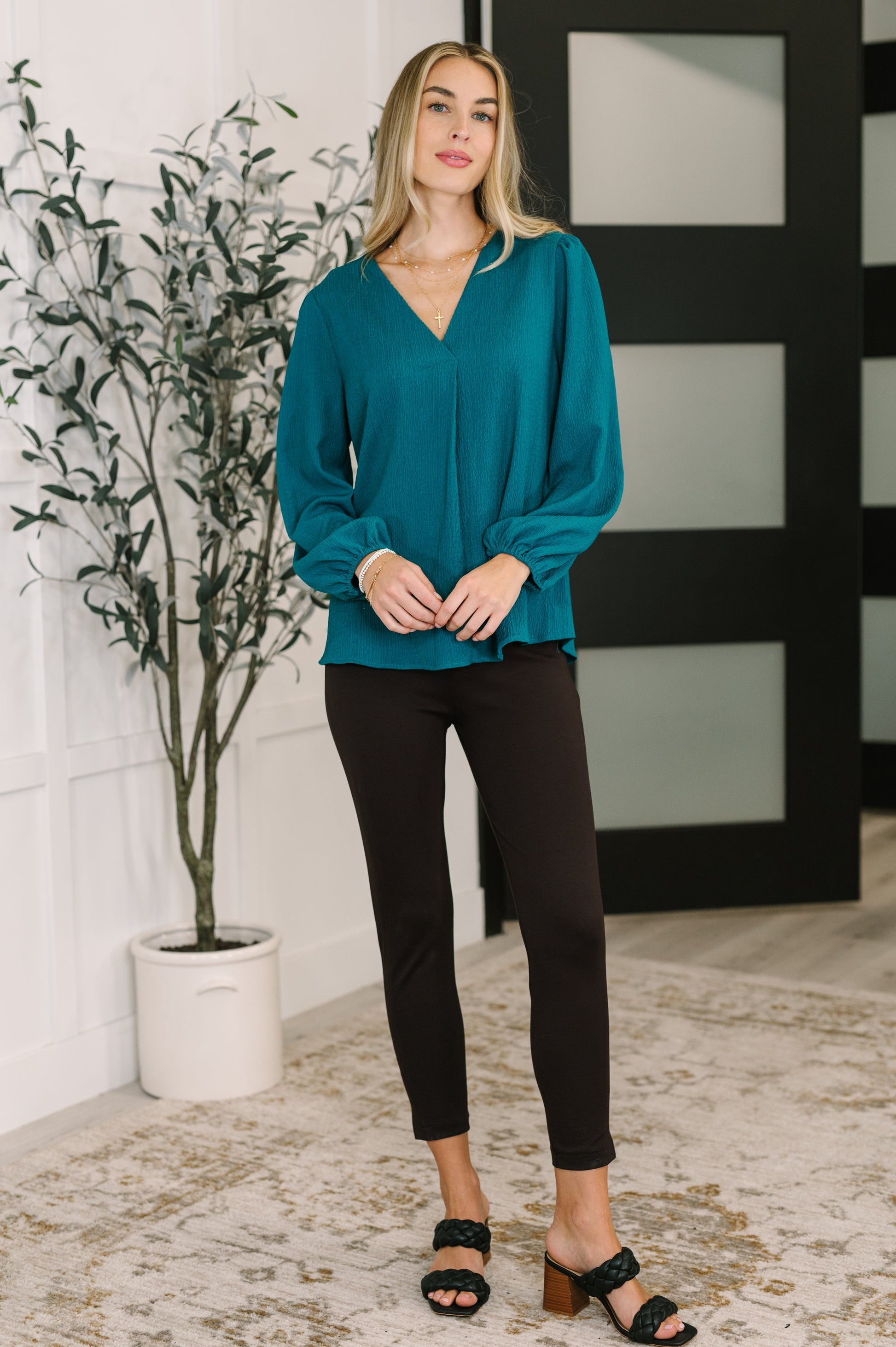 Woman wearing a teal blouse and black pants standing indoors with a plant and decorative wall in the background.