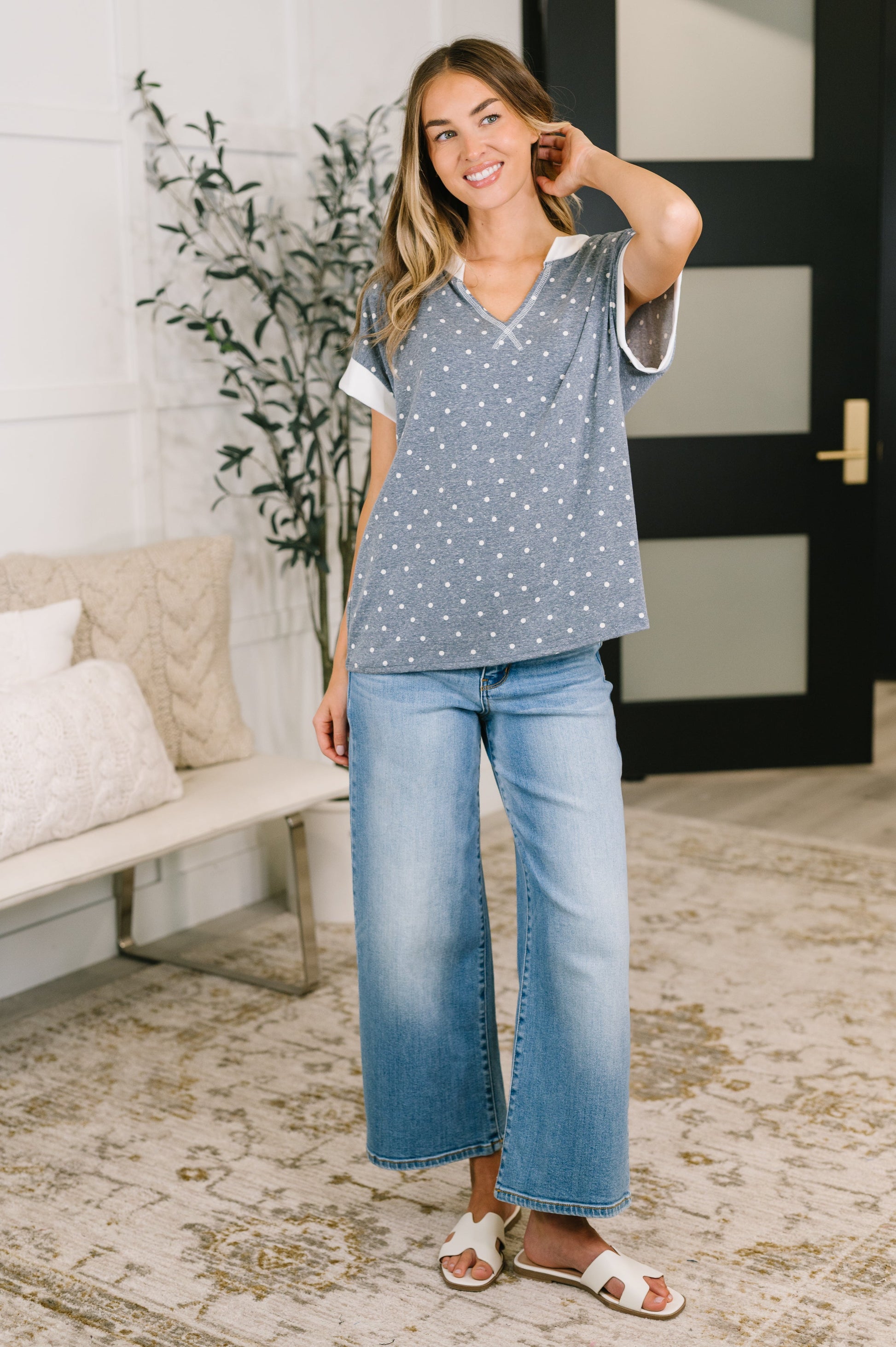 A woman modeling a blue short-sleeve top with white polka dots and light-wash wide-leg jeans in a bright, modern room setting.