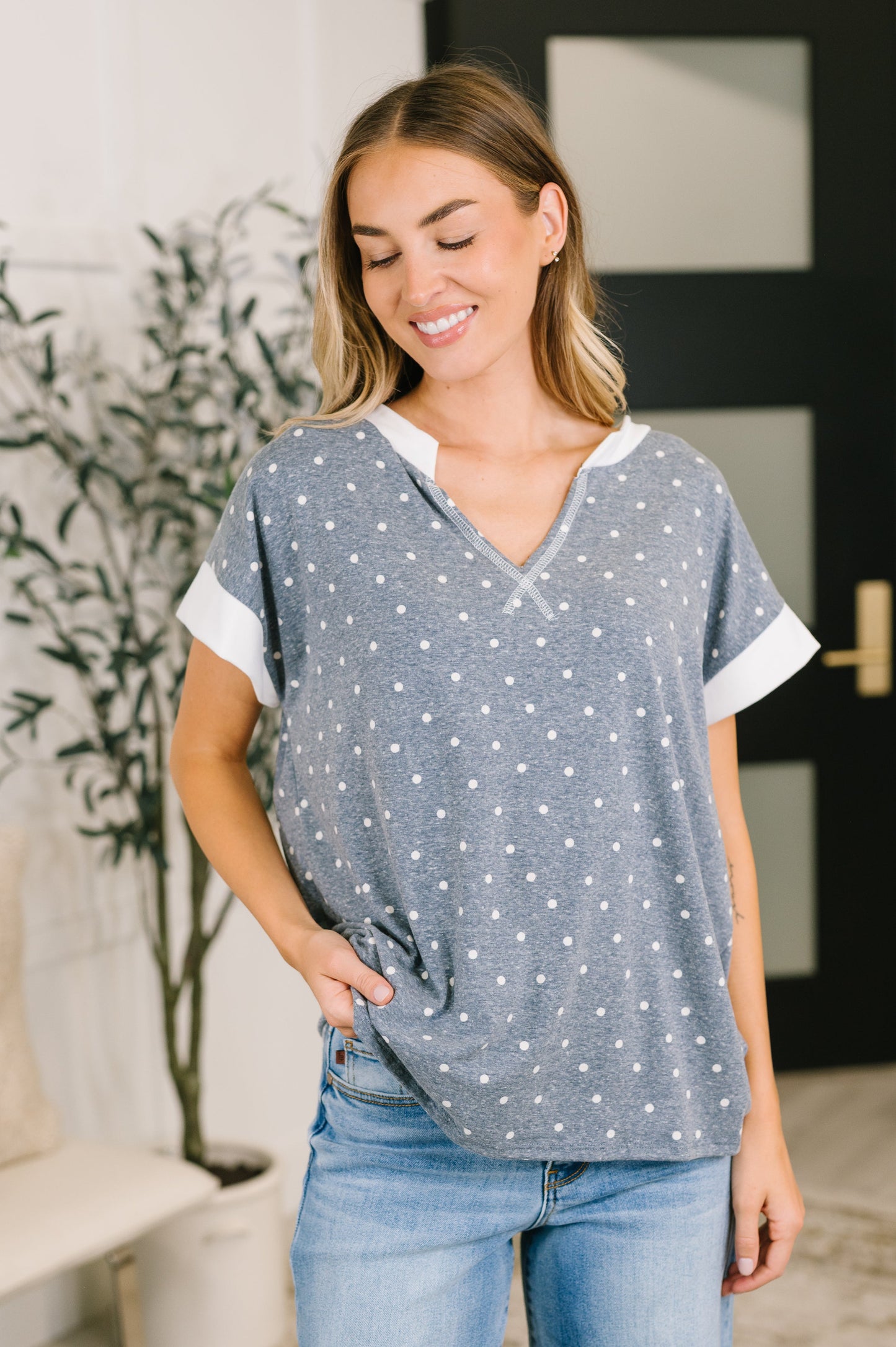 A woman wearing a gray short-sleeve top with white dots and white contrast trim, paired with blue jeans, standing in a brightly lit indoor setting.