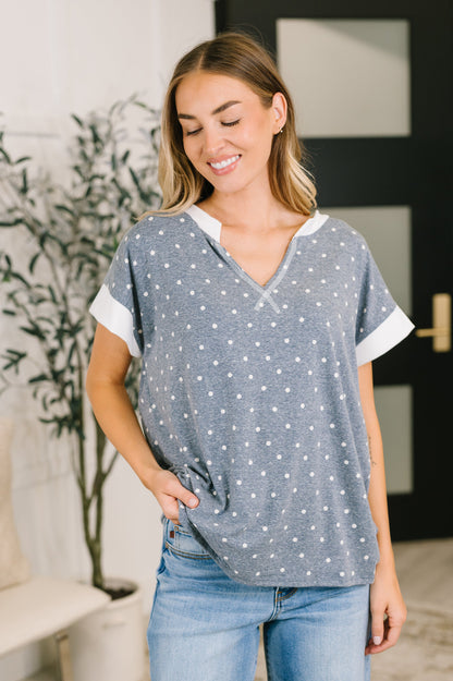 A woman wearing a gray short-sleeve top with white dots and white contrast trim, paired with blue jeans, standing in a brightly lit indoor setting.