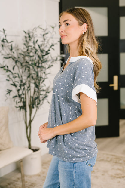 A woman wearing a gray short-sleeve top with white polka dots and white rolled cuffs, paired with light blue jeans, standing in a brightly lit indoor setting.