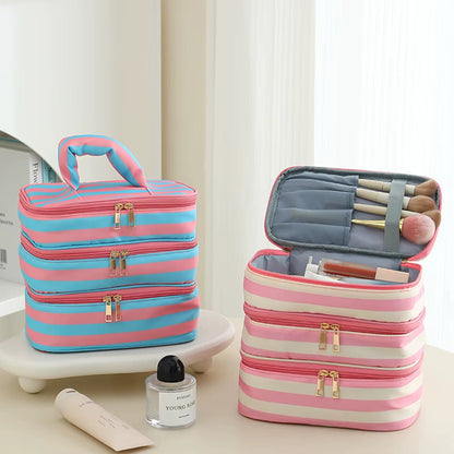 Two striped makeup bag sets are shown on a light beige surface with a white curtain background. One set is stacked closed in blue and pink, and the other is open showing makeup brushes and cosmetics inside, with a small perfume bottle and cream tube nearby as props.