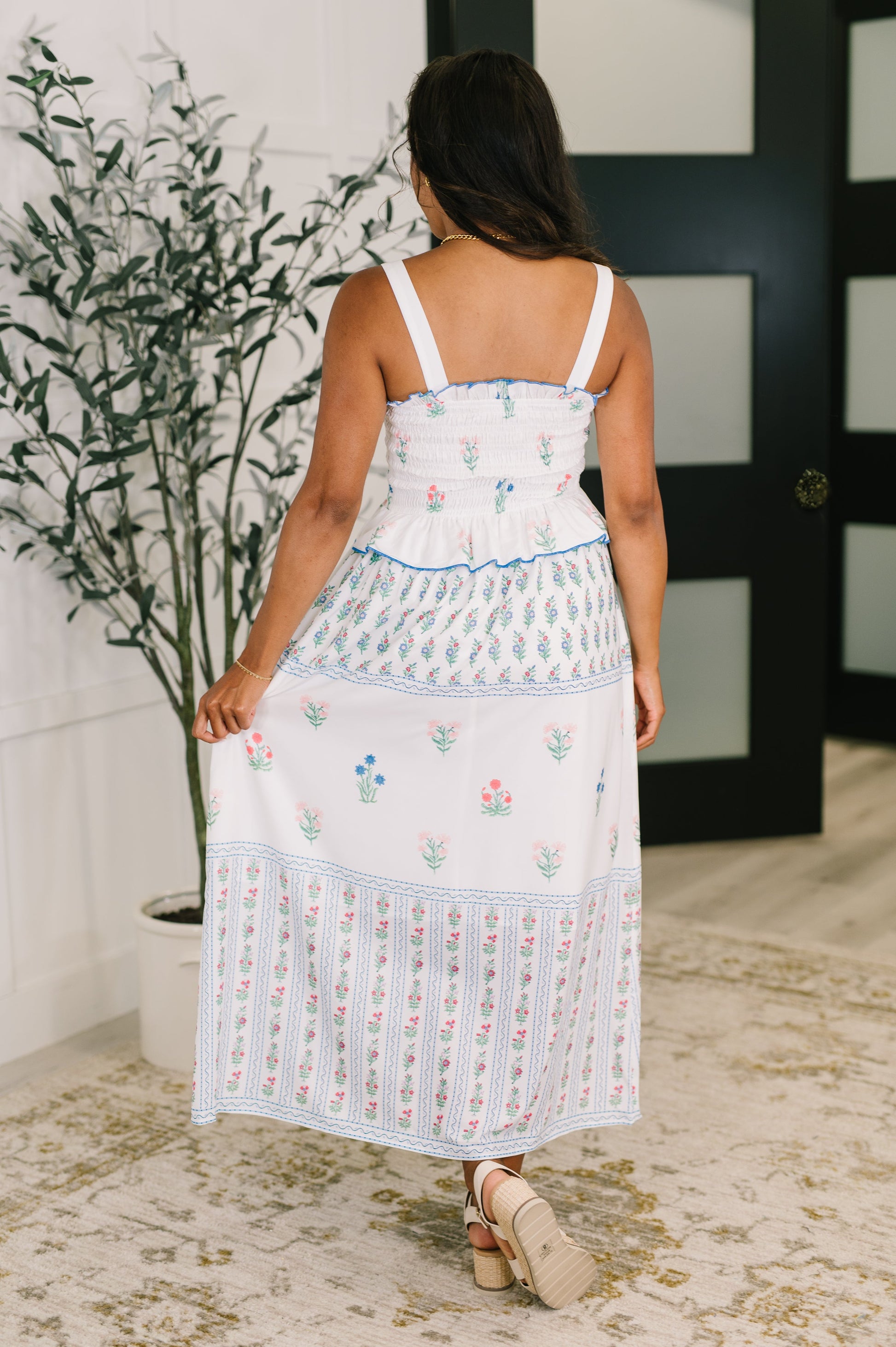 Woman wearing a white embroidered dress standing in a room with a plant and door in the background.