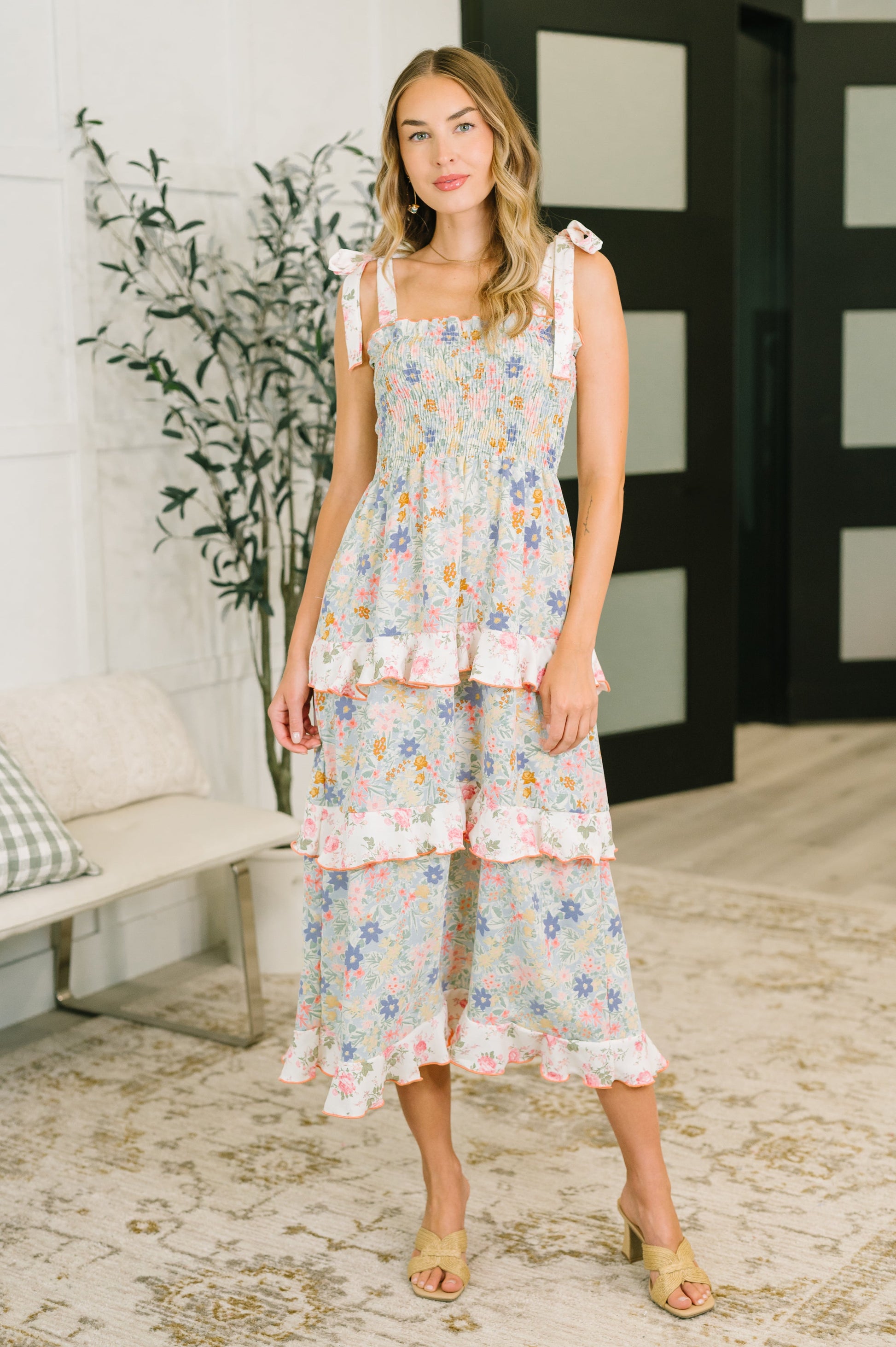 Woman wearing a floral dress standing in a room with a plant and bench.