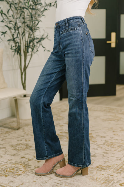 A person wearing dark blue high-waisted flare jeans, styled with a white top and pink block-heel mules, standing on a patterned rug in an indoor setting.