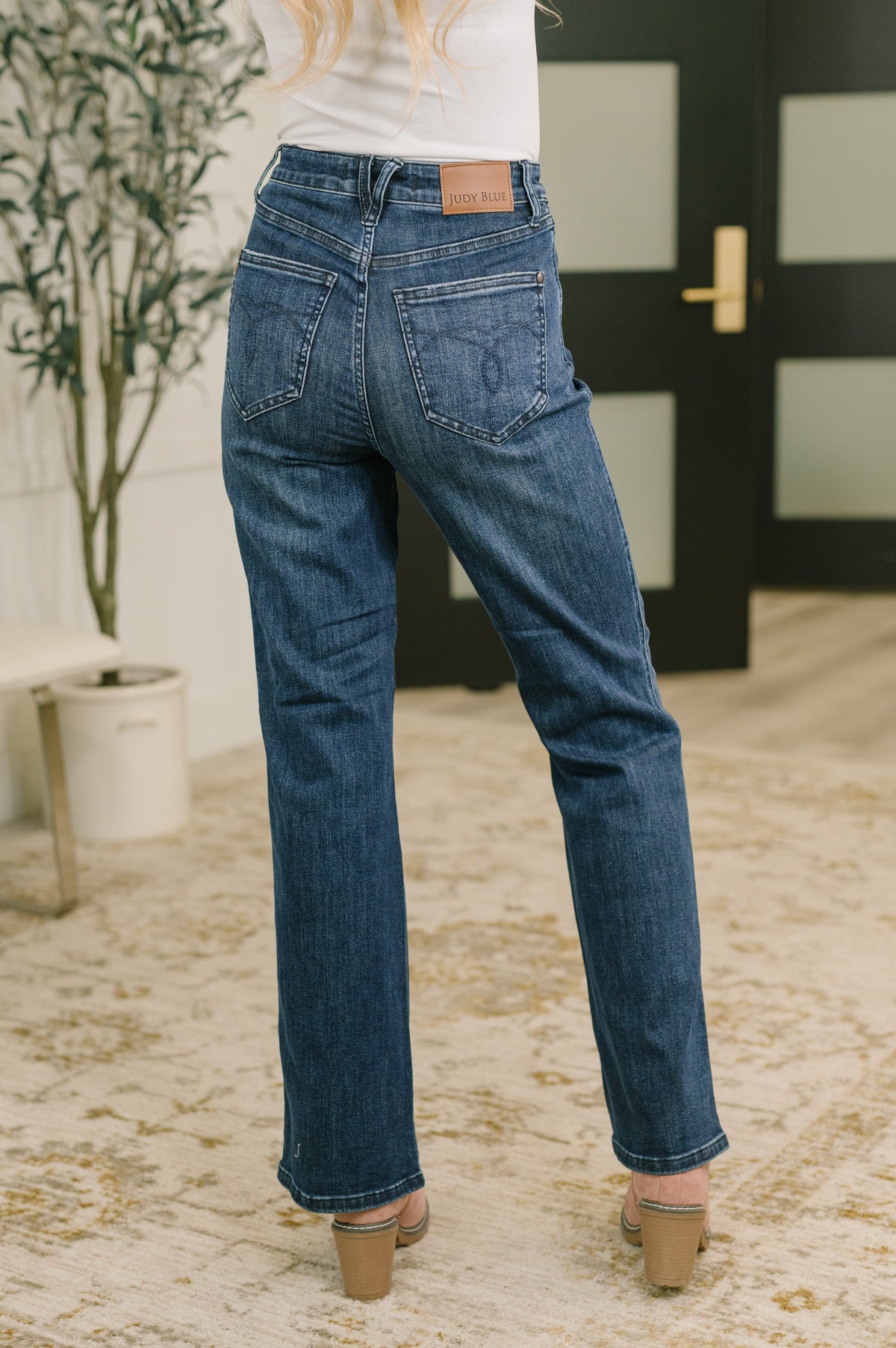 A person shown from the back wearing dark blue straight-leg jeans, a white top, and beige block-heel sandals in an indoor setting with a patterned rug and a potted plant.