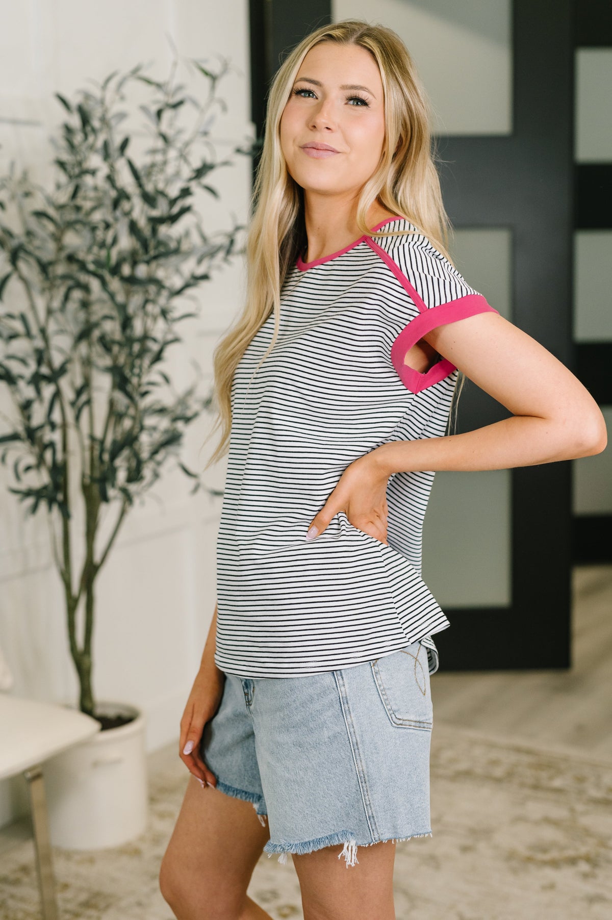 A woman modeling a short-sleeve ringer top with thin black and white horizontal stripes and bright pink trim, paired with light blue denim shorts in an indoor setting.