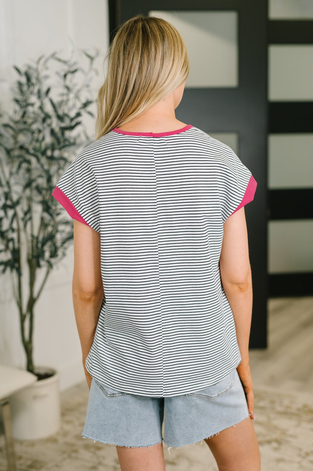 A woman viewed from behind wearing a gray and white striped t-shirt with bright pink trim on the collar and sleeve cuffs, paired with light blue denim shorts in an indoor setting.