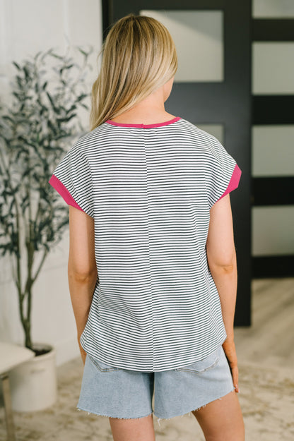A woman viewed from behind wearing a gray and white striped t-shirt with bright pink trim on the collar and sleeve cuffs, paired with light blue denim shorts in an indoor setting.
