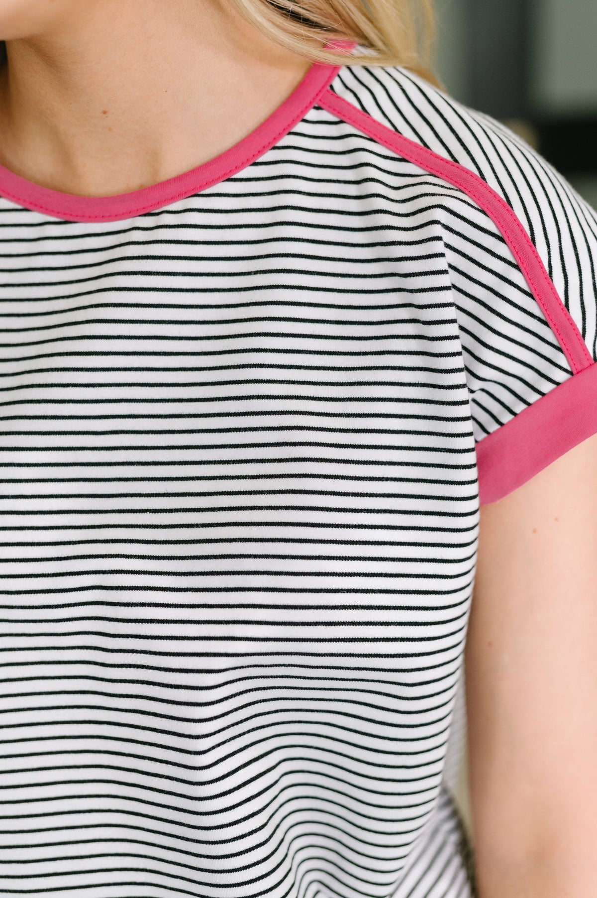 Close-up of a short-sleeve ringer T-shirt with thin black horizontal stripes on a white base and bright pink trim along the crew neck and sleeve cuffs, worn by a model.